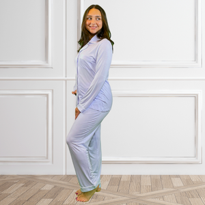 Variant::lilac  A young woman wearing a lilac bamboo long pj set with her hand on her hip, looking over her shoulder and smiling to the side.  The collar is a fold-down notched collar and the there is a breast pocket. The bamboo long pj set is trimmed in white piping.