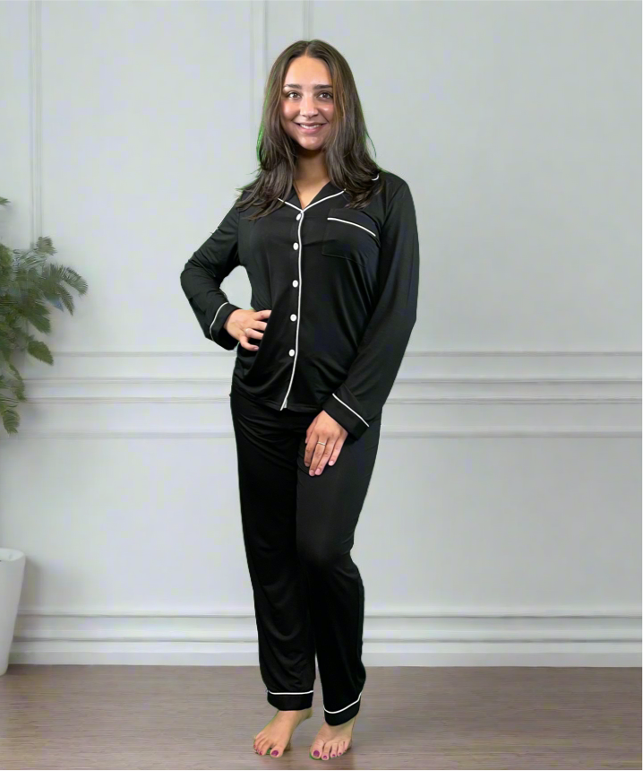 variant::black Woman wearing bamboo adaptive black long pj set. Has her hand on her hip and is smiling into the camera.  The collar is a fold-down notched collar and the there is a breast pocket. The adaptive long pj set is trimmed in white piping.