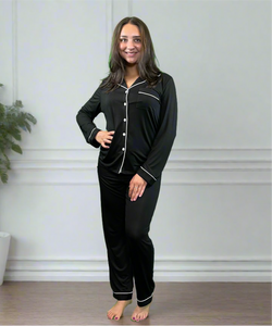 variant::black Woman wearing bamboo adaptive black long pj set. Has her hand on her hip and is smiling into the camera.  The collar is a fold-down notched collar and the there is a breast pocket. The adaptive long pj set is trimmed in white piping.