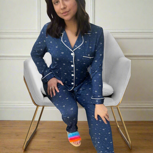 variant::blue-with-hearts A young woman with dark hair wearing navy heart adaptive bamboo long pajama set, while sitting on a chair. The collar is a fold-down notched collar and there is a breast pocket. The adaptive pj set is trimmed in white piping.