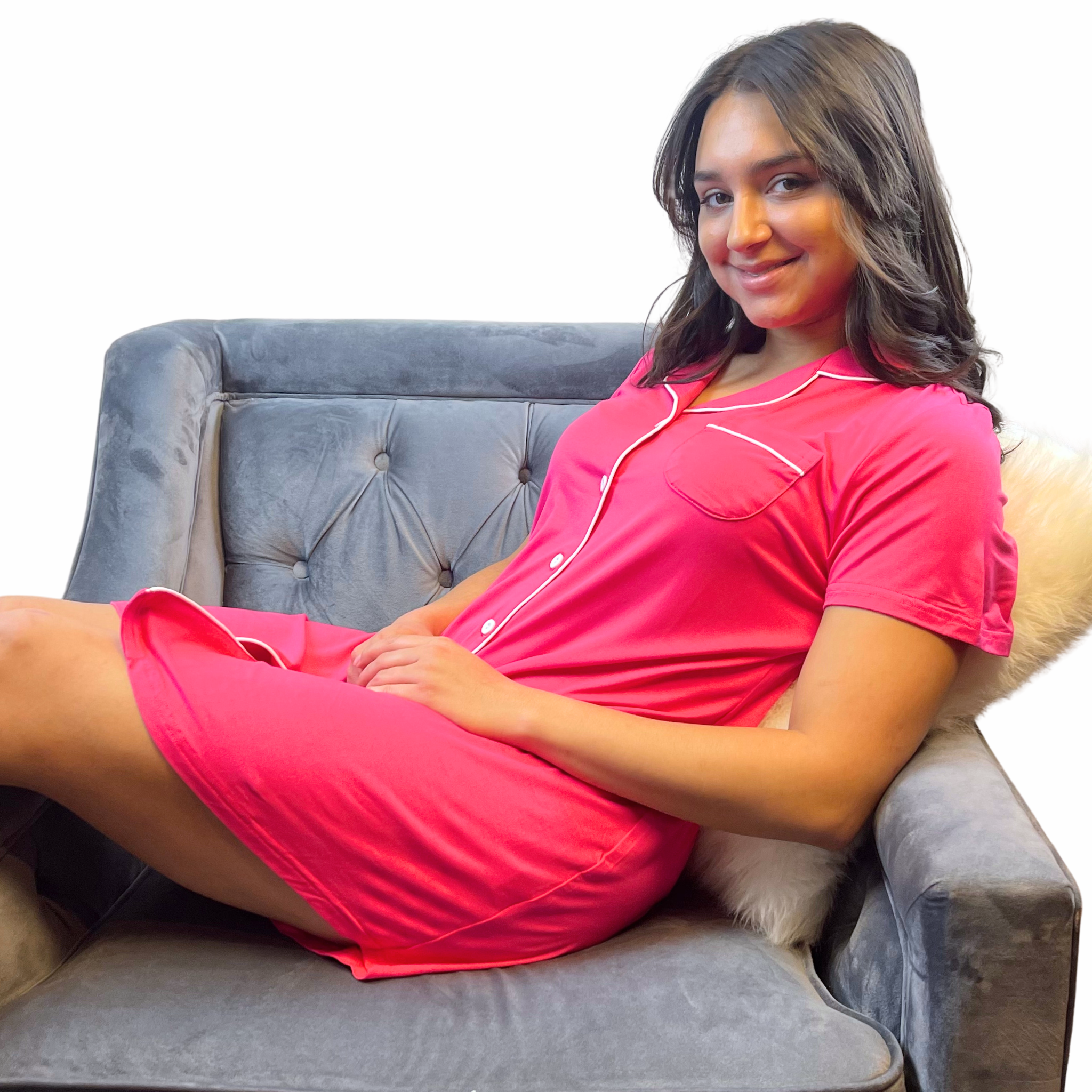 variant:fuchsia A young woman with long, dark hair wearing a bright hot pink adaptive nightgown with decorative buttons and Velcro® closures. She is sitting across an upholstered gray chair with her legs hanging over the side. The collar is a fold-down notched collar and the there is a breast pocket. The adaptive nightgown is trimmed in white piping. She had her hands on her lap and is smiling. 