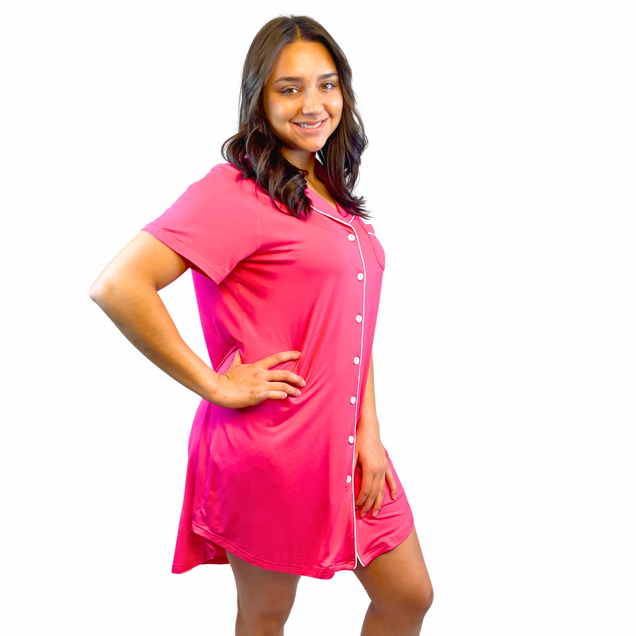variant:fuchsia A young woman with long, dark hair wearing a bright hot pink adaptive nightgown with decorative buttons and Velcro® closures. The photo is from the side and she has one hand on her hip and looking at the camera smiling. The collar is a fold-down notched collar and the there is a breast pocket. The adaptive nightgown is trimmed in white piping. 