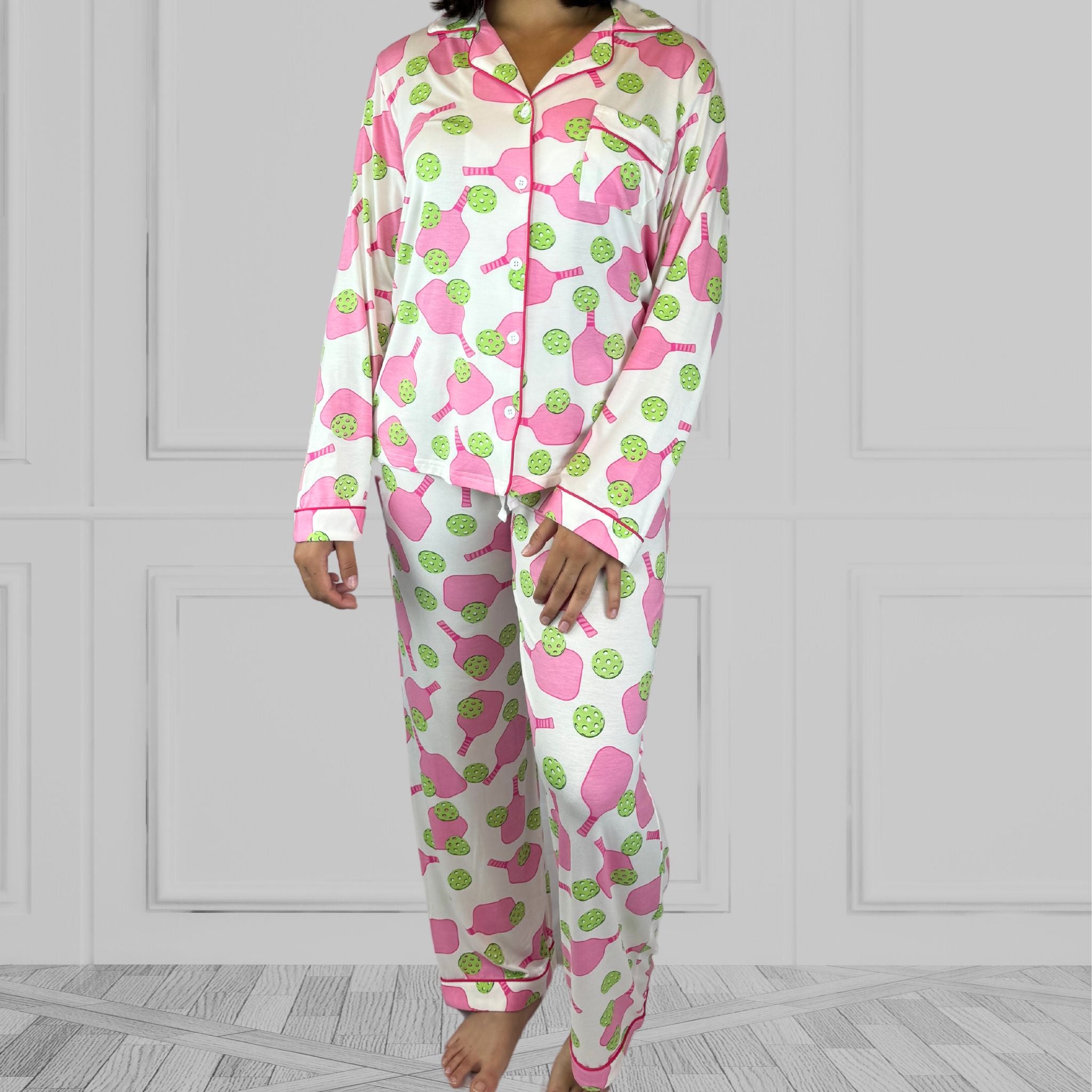 variant::preppy-pickleball A full view of a young woman with dark hair wearing pink and green pickleball printed pajamas with one hand on her hip.  The collar is a fold-down notched collar and the there is a breast pocket. The bamboo long sleeve pajama set is trimmed in pink piping.