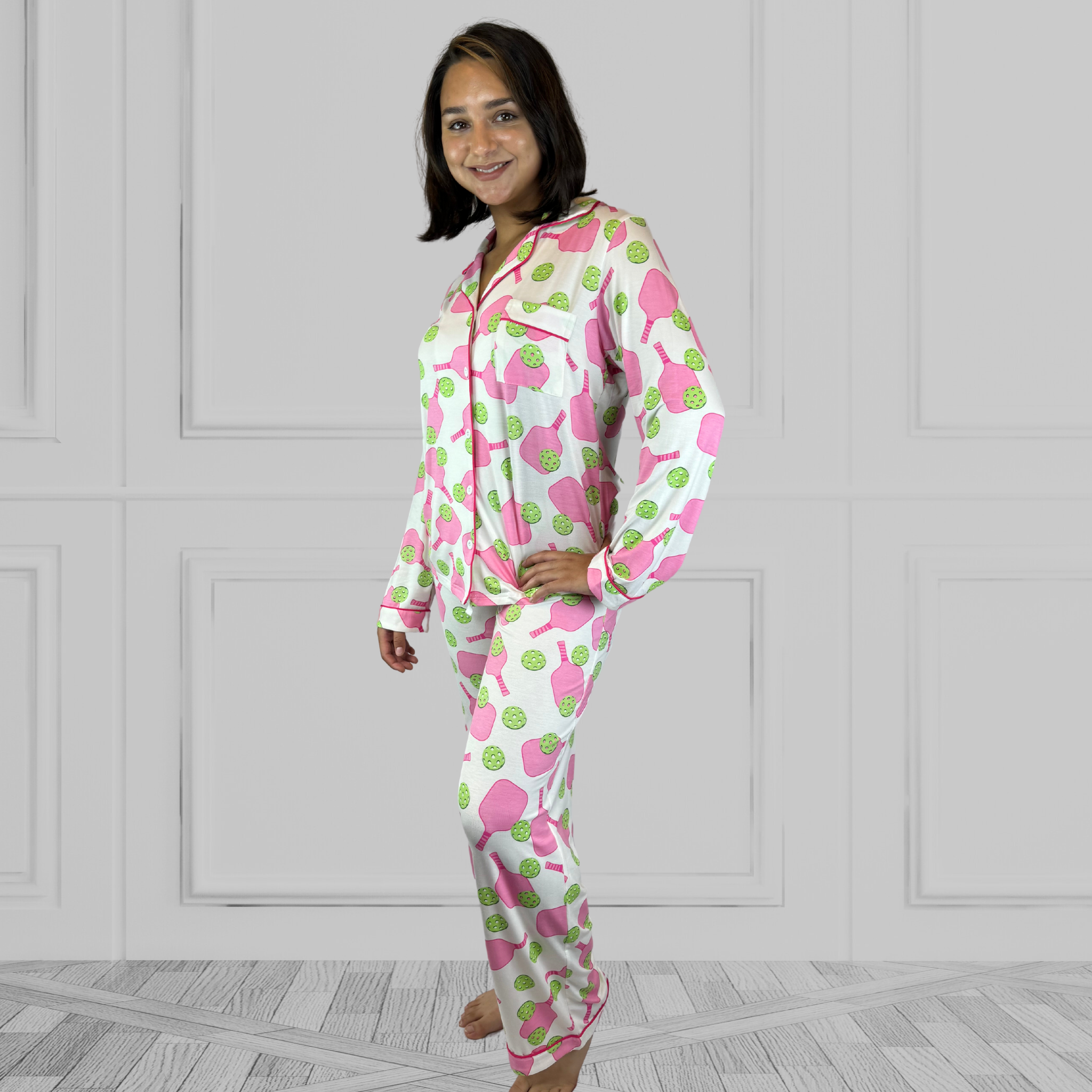 variant::preppy-pickleball A full view of a young woman with dark hair wearing pink and green pickleball printed pajamas with one hand on her hip.  The collar is a fold-down notched collar and the there is a breast pocket. The bamboo long sleeve pajama set is trimmed in pink piping.