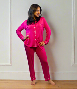 variant::fuchsia Woman wearing fuchsia long sleeve bamboo adaptive pajamas. She has both hands on her hips and turned to the side, looking into the distance and smiling.  The collar is a fold-down notched collar and the there is a breast pocket. The adaptive long pj set is trimmed in white piping.