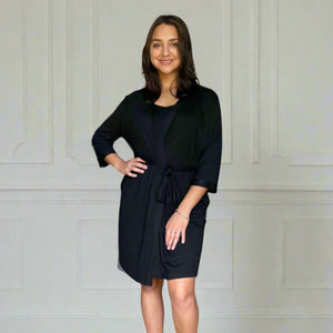 variant::black Women's Bamboo Robe modeled by a young woman with shoulder-length brown hair. She is turned to the camera with one hand on her hip, smiling. The bamboo robe is black and has a v-neck with pockets for functionality. The sleeves are three quarter sleeves and the robe falls right above her knee.