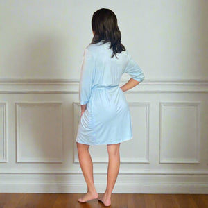 variant::light blue Women's Bamboo Robe modeled by a young woman with shoulder-length brown hair. She is turned to the wall, showcasing the back of the light blue, bamboo robe. The robe is v-neck, has three quarters sleeves, and the robe falls just above her knee