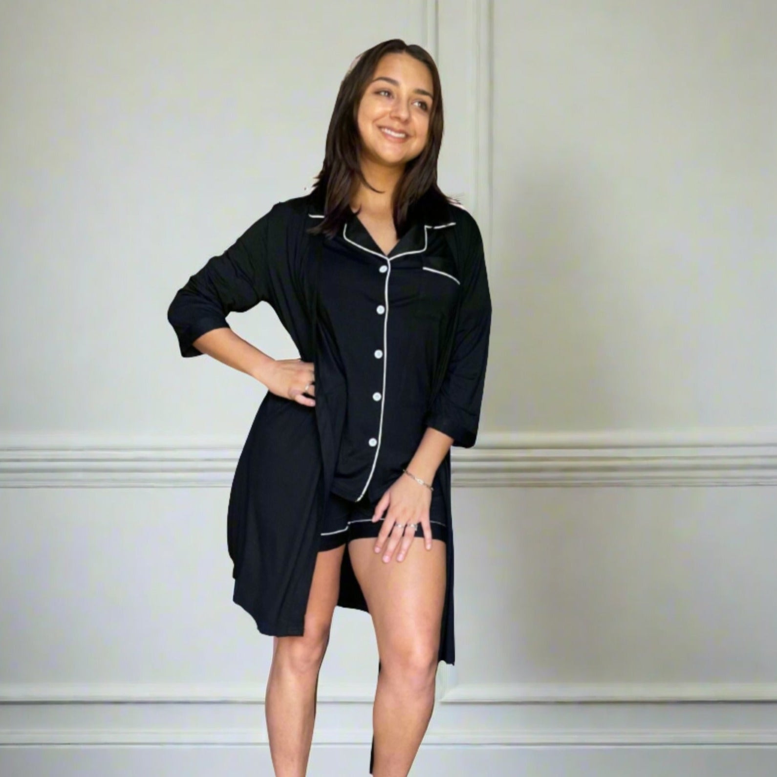 variant::black Women's Bamboo Robe modeled by a young woman with shoulder-length brown hair. She is turned to the camera with one hand on her hip, smiling off into the distance. The bamboo robe is black and has a v-neck with pockets for functionality. The sleeves are three quarter sleeves and the robe falls right above her knee.