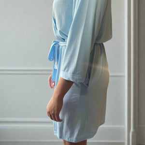 variant::light blue Women's Bamboo Robe modeled by a young woman with shoulder-length brown hair. She is turned to the side, showcasing the sleeves of the light blue, bamboo robe. The robe is v-neck, has three quarters sleeves, and the robe falls just above her knee.