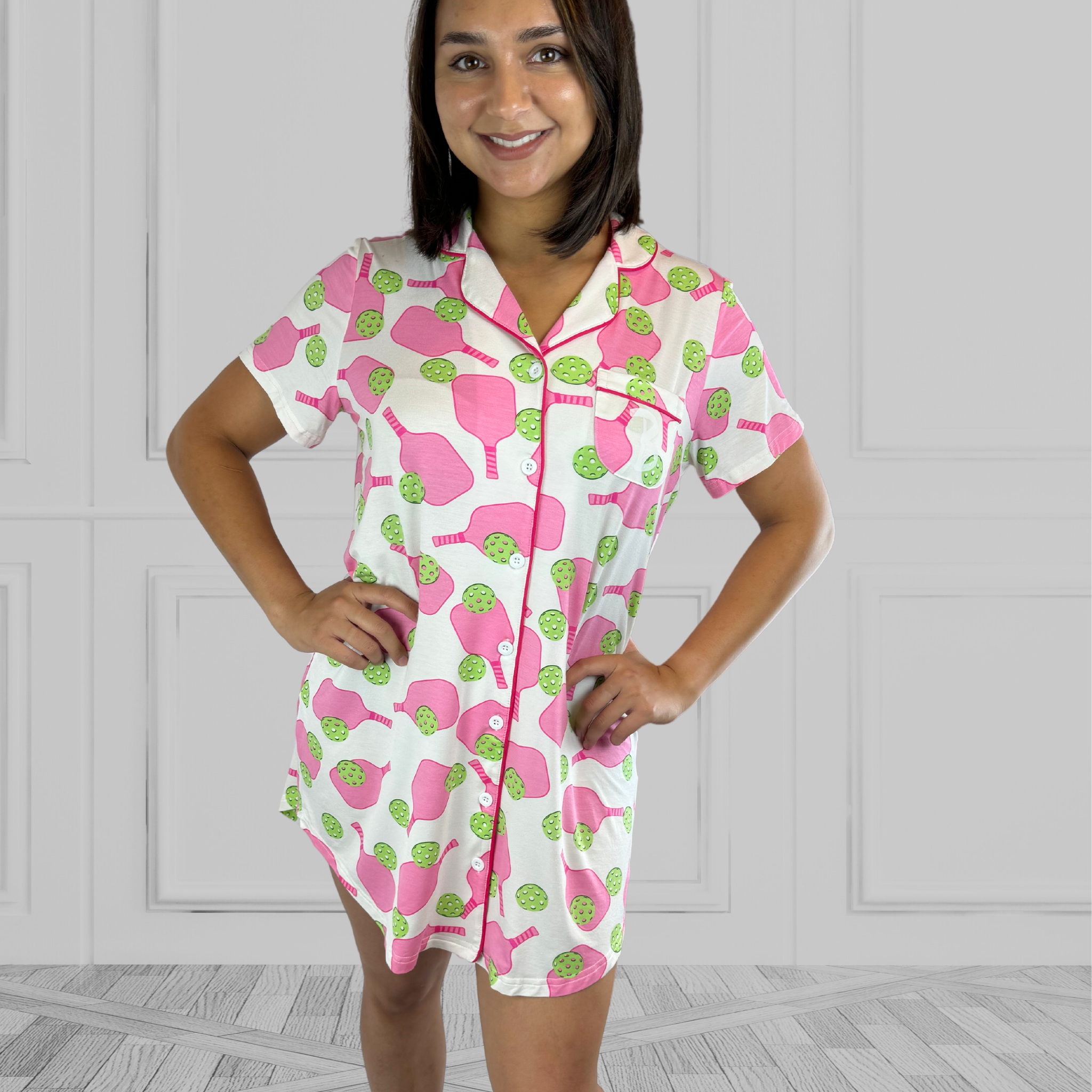 variant:pickleball print Pink and green bamboo pickleball print nightgown worn by young woman with dark hair facing the camera with her hands on her hips. The nightgown has decorative buttons up the front and is trimmed in hot pink piping. The closure under the buttons is Velcro®. The hot pink paddles and bright green balls are on a crisp white background. The sleeves are short and the longest part of the shirttail hem comes  3 inches above the knee. The shortest part of the hem comes above her mid-thigh. 