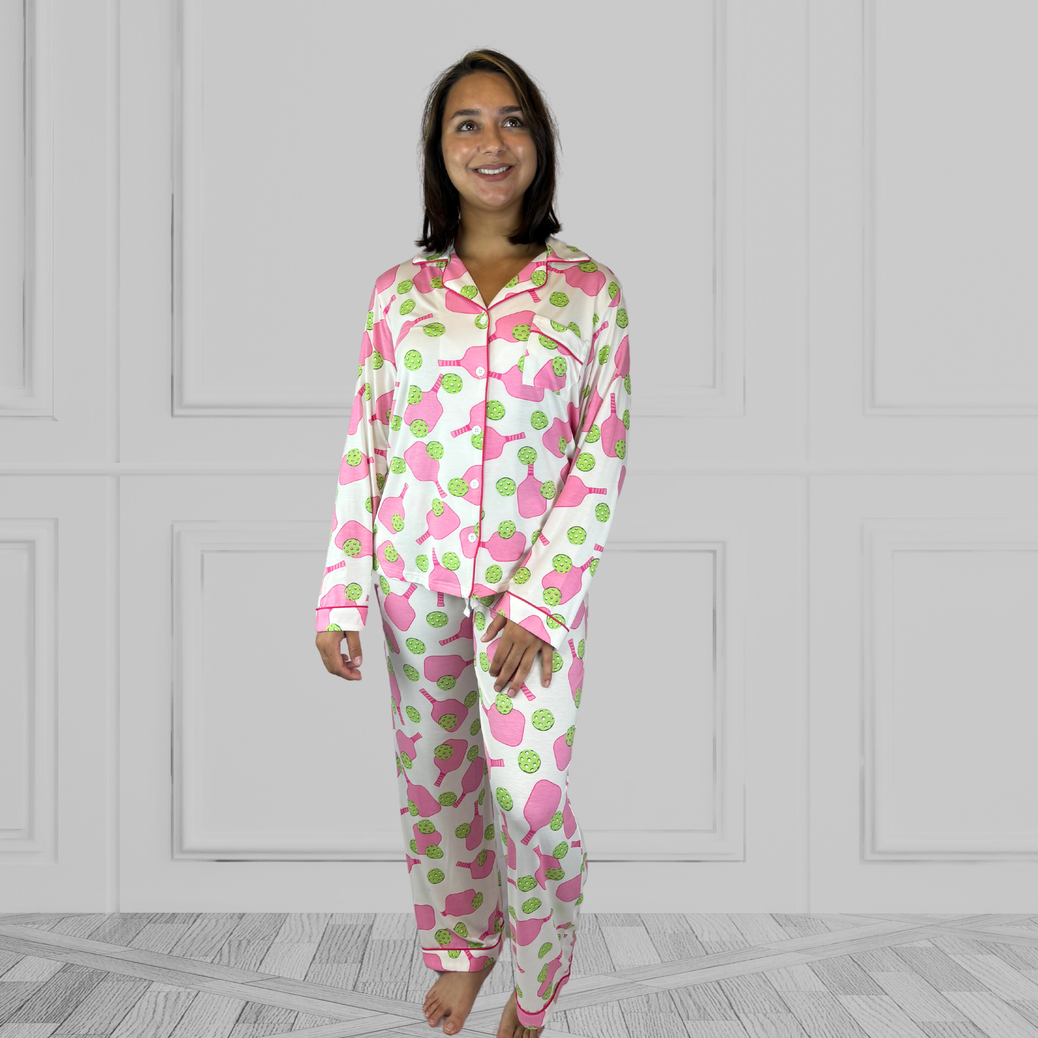 variant::preppy-pickleball A full view of a young woman with dark hair wearing pink and green pickleball printed pajamas with one hand on her hip.  The collar is a fold-down notched collar and the there is a breast pocket. The bamboo long sleeve pajama set is trimmed in pink piping.