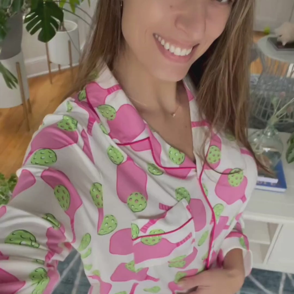 variant::preppy-pickleball A front view of a  young woman with dark hair wearing pink and green pickleball printed pajamas with one hand on her hip.  The collar is a fold-down notched collar and the there is a breast pocket. The bamboo long sleeve pajama set is trimmed in pink piping.