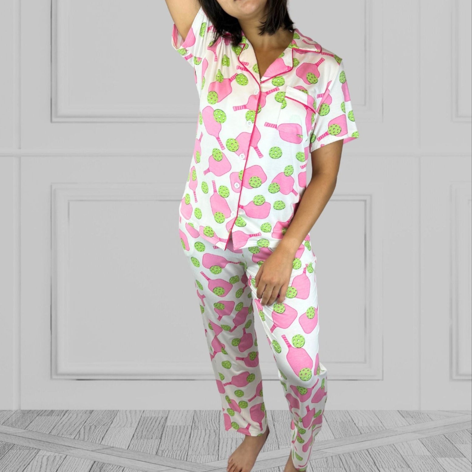 variant::preppy-pickleball Pink and green bamboo pickleball pajama set worn by young woman with dark hair. and one arm above her head. The background of the pajams is crisp white and the sleeves are short. The pants are cropped jogger style with a tapered leg. 