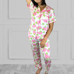 variant::preppy-pickleball Pink and green bamboo pickleball pajama set worn by young woman with dark hair. and one arm above her head. The background of the pajams is crisp white and the sleeves are short. The pants are cropped jogger style with a tapered leg. 