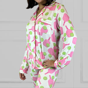 variant::preppy-pickleball A young woman with dark hair wearing pink and green pickleball printed pajamas with one hand on her hip.  The collar is a fold-down notched collar and the there is a breast pocket. The bamboo long sleeve pajama set is trimmed in pink piping.