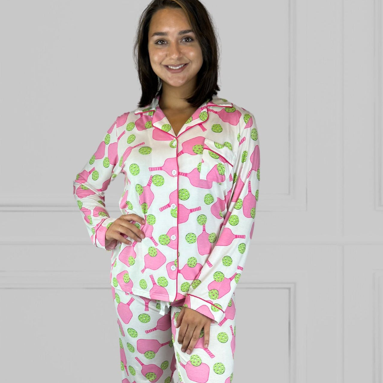 variant::preppy-pickleball A front view of a  young woman with dark hair wearing pink and green pickleball printed pajamas with one hand on her hip.  The collar is a fold-down notched collar and the there is a breast pocket. The bamboo long sleeve pajama set is trimmed in pink piping.