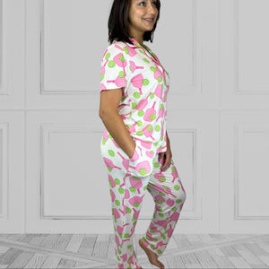 variant::preppy-pickleball Pink and green bamboo pickleball pajama set worn by young woman with dark hair. standing at a side view with one hand in her pocket. The background of the pajams is crisp white and the sleeves are short. The pants are cropped jogger style with a tapered leg. 