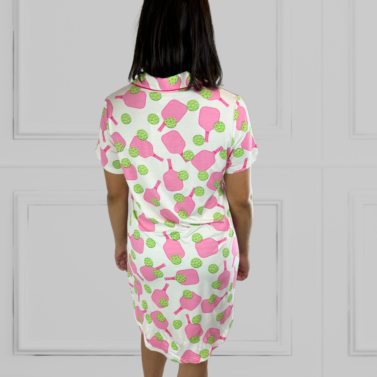 variant::preppy-pickleball Pink and green bamboo pickleball print nightgown worn by young woman with dark hair with her back to the camera. Her hands are to her side. The hot pink paddles and bright green balls are on a crisp white background. The sleeves are short and the longest part of the rear shirttail hem comes  to her knee. The shortest part of 3the hem comes above her mid-thigh. 