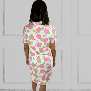variant:pickleball print Back view of pink and green bamboo pickleball print nightgown worn by young woman with dark hair facing the camera with her hands on her hips. The nightgown has decorative buttons up the front and is trimmed in hot pink piping. The closure under the buttons is Velcro®. The hot pink paddles and bright green balls are on a crisp white background. The sleeves are short and the longest part of the shirttail hem comes  3 inches above the knee.
