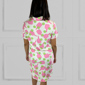 variant::preppy-pickleball Pink and green bamboo pickleball print nightgown worn by young woman with dark hair with her back to the camera. Her hands are to her side. The hot pink paddles and bright green balls are on a crisp white background. The sleeves are short and the longest part of the rear shirttail hem comes  to her knee. The shortest part of 3the hem comes above her mid-thigh. 