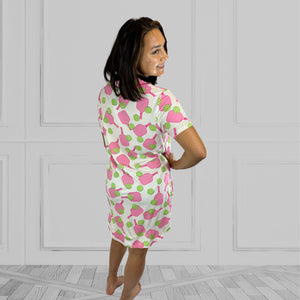 variant::preppy-pickleball Pink and green bamboo pickleball print nightgown worn by young woman with dark hair with her back to the camera with one hand on her hip. The hot pink paddles and bright green balls are on a crisp white background. The sleeves are short and the longest part of the shirttail hem comes  3 inches above the knee. The shortest part of the hem comes above her mid-thigh. The nightown also has a notched collar and breast pocket. 