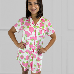 variant::preppy-pickleball Pink and green bamboo pickleball print nightgown worn by young woman with dark hair facing the camera with her hands on her hips. The nightgown buttons up the front and is trimmed in hot pink piping. The hot pink paddles and bright green balls are on a crisp white background. The sleeves are short and the longest part of the shirttail hem comes  3 inches above the knee. The shortest part of the hem comes above her mid-thigh. It also has a notched collar and breast pocket. 