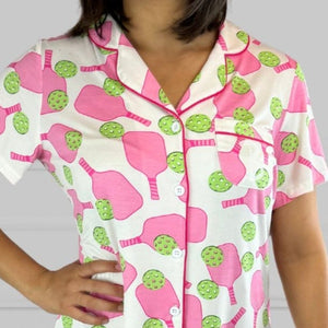 variant:preppy-pickeball  Closeup of pink and green bamboo pickleball print nightgown worn by young woman with dark hair facing the camera with her hands on her hips. The nightgown has decorative buttons up the front and is trimmed in hot pink piping. The hot pink paddles and bright green balls are on a crisp white background. The sleeves are short and the longest part of the shirttail hem comes 3 inches above the knee. The shortest part of the hem comes above her mid-thigh. 