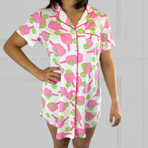 variant:pickleball print Pink and green adaptive bamboo pickleball print nightgown worn by young woman with dark hair facing the camera with her hands on her hips. Her head is not in the shot. The nightgown has decorative buttons up the front and is trimmed in hot pink piping. The closure under the buttons is Velcro®. The hot pink paddles and bright green balls are on a crisp white background. The hem comes above her mid-thigh. 