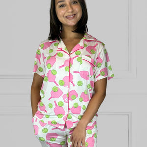variant::preppy-pickleball Pink and green bamboo pickleball pajama set worn by young woman with dark hair. and one hand in her pocket The background of the pajams is crisp white and the sleeves are short. The pants are cropped jogger style with a tapered leg. 