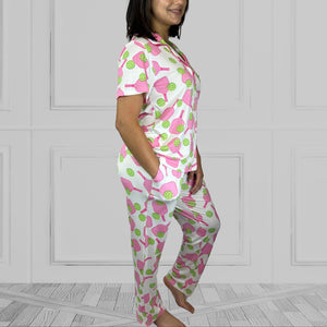 variant::preppy-pickleball Pink and green bamboo pickleball pajama set worn by young woman with dark hair. and one hand in her pocket standing at a side view. The background of the pajams is crisp white and the sleeves are short. The pants are cropped jogger style with a tapered leg.
