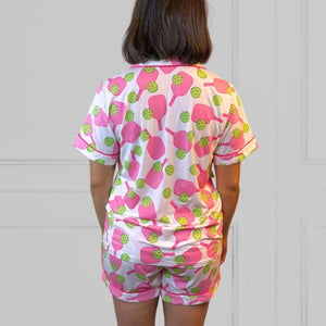 Back view of a young woman wearing a pink and green pickleball printed pajama shorty set.  The collar is a fold-down notched collar and the there is a breast pocket. The bamboo short set is trimmed in pink piping. There are short sleeves and the shorts come to about mid-thigh.