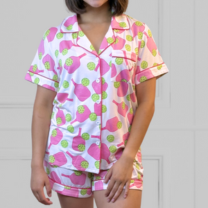 a young woman wearing a pink and green pickleball printed pajama shorty set.  The collar is a fold-down notched collar and the there is a breast pocket. The bamboo short set is trimmed in pink piping. There are short sleeves and the shorts come to about mid-thigh.