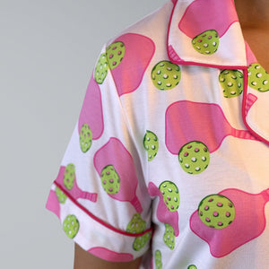 variant::preppy-pickleball Close-up, shoulder-only pink and green bamboo pickleball print nightgown worn by young woman. Shownn is the shoulder, sleeve and collar.  The nightgown is trimmed in hot pink piping. The hot pink paddles and bright green balls are on a crisp white background. The sleeves are short.  