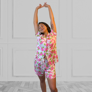 a young woman wearing a pink and green pickleball printed pajama shorty set.  The collar is a fold-down notched collar and the there is a breast pocket. The bamboo short set is trimmed in pink piping. There are short sleeves and the shorts come to about mid-thigh.