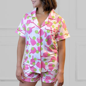 a young woman wearing a pink and green pickleball printed pajama shorty set.  The collar is a fold-down notched collar and the there is a breast pocket. The bamboo short set is trimmed in pink piping. There are short sleeves and the shorts come to about mid-thigh.